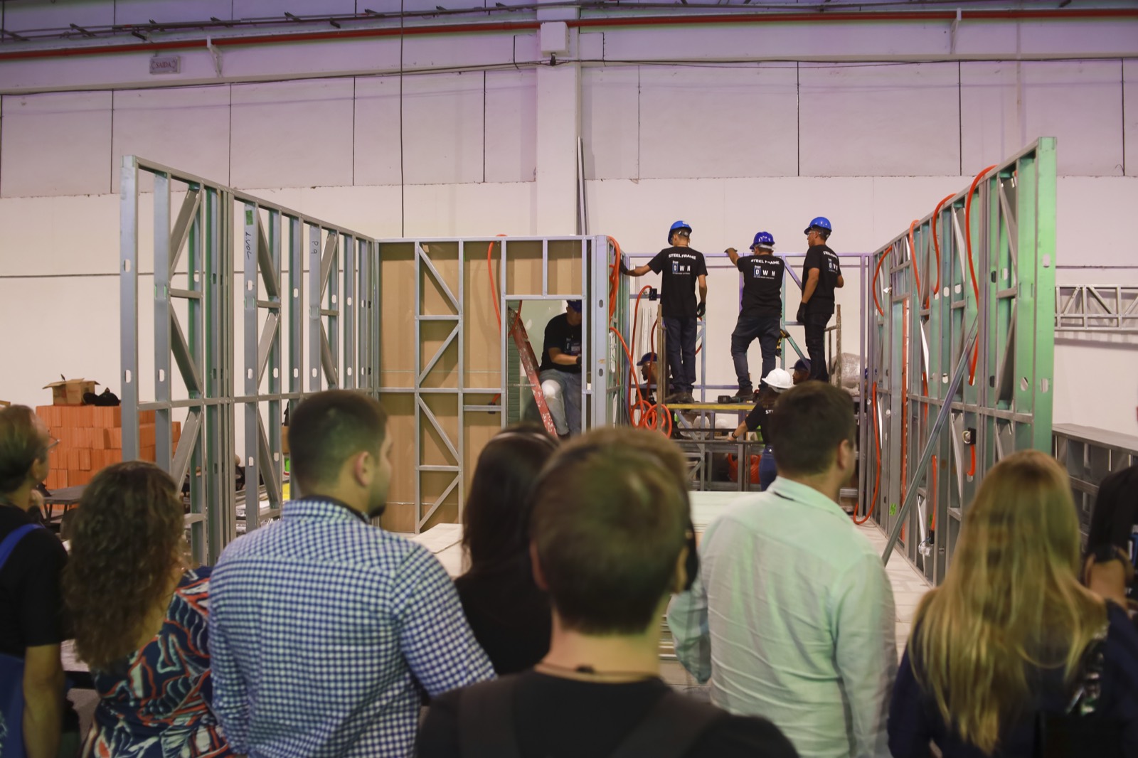 Demonstração de montagem em light steel frame, com montadores trabalhando e público assistindo